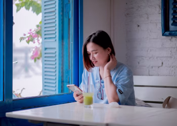 A young woman in a light blue shirt checks her smartphone while enjoying a drink by a window in a quaint cafe.
