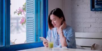 A young woman in a light blue shirt checks her smartphone while enjoying a drink by a window in a quaint cafe.