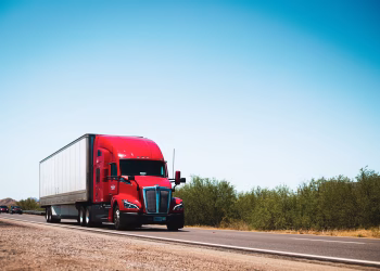 Vibrant red semi-trailer truck driving on an open highway under a clear blue sky.