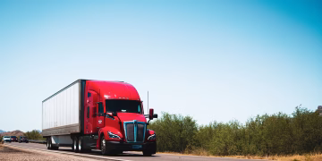 Vibrant red semi-trailer truck driving on an open highway under a clear blue sky.