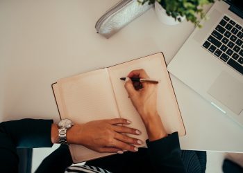 Person writing in notebook while using laptop at a modern workspace