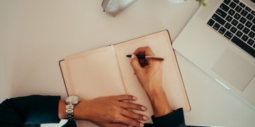 Person writing in notebook while using laptop at a modern workspace