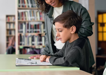 A young boy engages with a book in a library under guidance, fostering a love for learning.