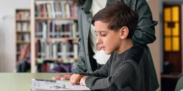 A young boy engages with a book in a library under guidance, fostering a love for learning.