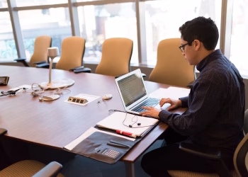A professional adult sitting at a desk with a laptop in a bright, modern conference room.