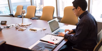 A professional adult sitting at a desk with a laptop in a bright, modern conference room.