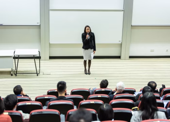 Female speaker presenting to an audience in a modern auditorium setting.