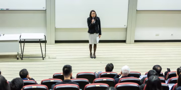 Female speaker presenting to an audience in a modern auditorium setting.