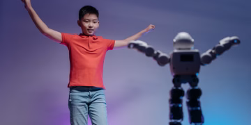 Asian boy in red polo shirt mimicking robot's dance moves under colorful lighting indoors.