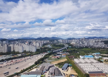 Aerial panorama of Guri City with urban skyline, highways, and mountains under a cloudy sky.
