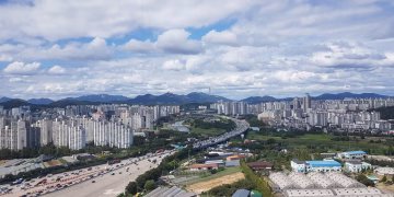 Aerial panorama of Guri City with urban skyline, highways, and mountains under a cloudy sky.