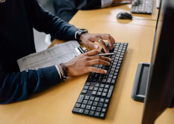 Close-up of hands typing on a keyboard in an office setting with clipboard nearby.