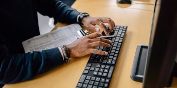 Close-up of hands typing on a keyboard in an office setting with clipboard nearby.