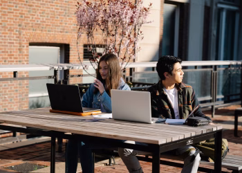 Two students studying outdoors with laptops on a university campus during the day.