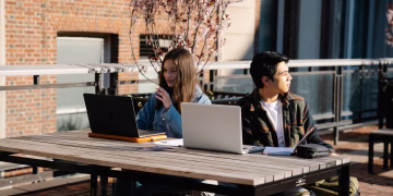 Two students studying outdoors with laptops on a university campus during the day.