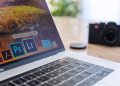 Close-up of a laptop displaying Adobe software icons, beside a camera on a desk.