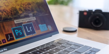 Close-up of a laptop displaying Adobe software icons, beside a camera on a desk.