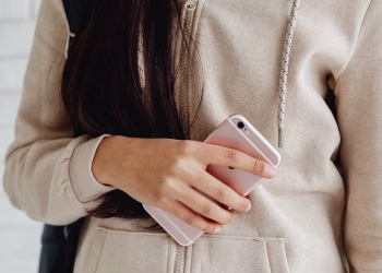 Young woman in a hoodie holding a pink smartphone indoors. Casual and modern lifestyle.