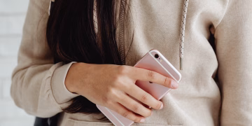 Young woman in a hoodie holding a pink smartphone indoors. Casual and modern lifestyle.