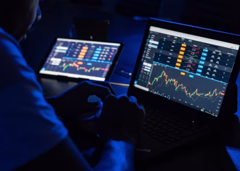 Man analyzing financial charts and data on laptops in a dimly lit room, highlighting forex trading.