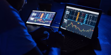 Man analyzing financial charts and data on laptops in a dimly lit room, highlighting forex trading.