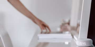 A smart speaker with a digital clock display placed on a minimalist desk setting, hand in background.