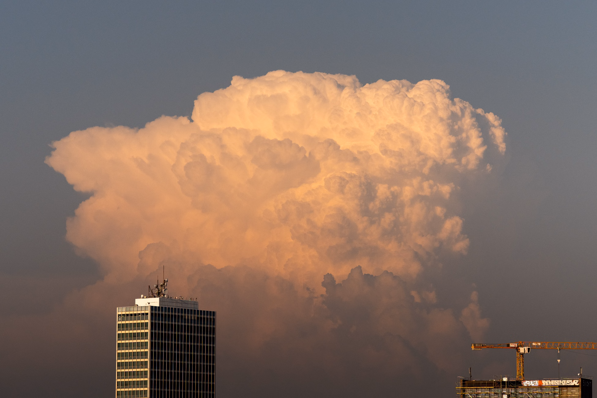 A towering cumulonimbus cloud illuminated by sunset over buildings in Düsseldorf, symbolizing urban nature contrast.