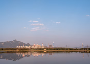 Scenic view of a power plant by a lake, reflecting mountains and a sky with scattered clouds.