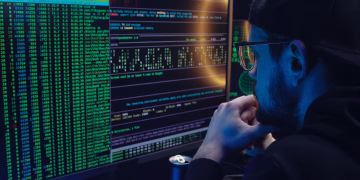 Man intently working on computer programming with code displayed on dual monitors in a dimly lit room.