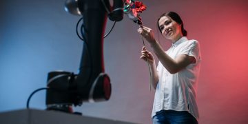 A woman and a robot arm collaboratively holding a flower, symbolizing technology's gentle touch.