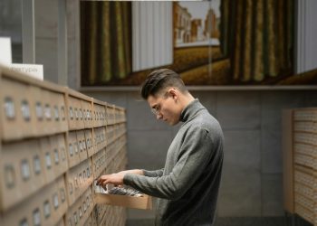A man stands in a library looking through card files from a drawer, surrounded by bookshelves.