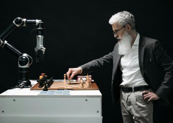 A bearded man engages in a chess game with a robotic arm, highlighting technology and strategy.