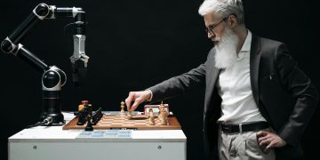 A bearded man engages in a chess game with a robotic arm, highlighting technology and strategy.