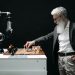 A bearded man engages in a chess game with a robotic arm, highlighting technology and strategy.