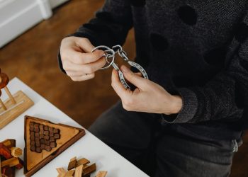 Top view of hands solving metal puzzle, surrounded by wooden brain teasers on a table.