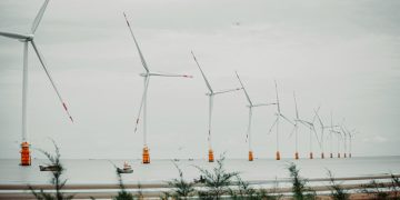 Scenic view of offshore wind turbines lined up in calm sea setting with vegetation in foreground.