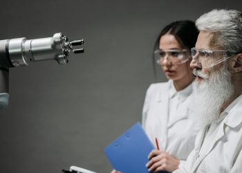 Two scientists wearing lab coats and goggles analyzing a robotic arm in a laboratory setting.