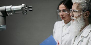 Two scientists wearing lab coats and goggles analyzing a robotic arm in a laboratory setting.