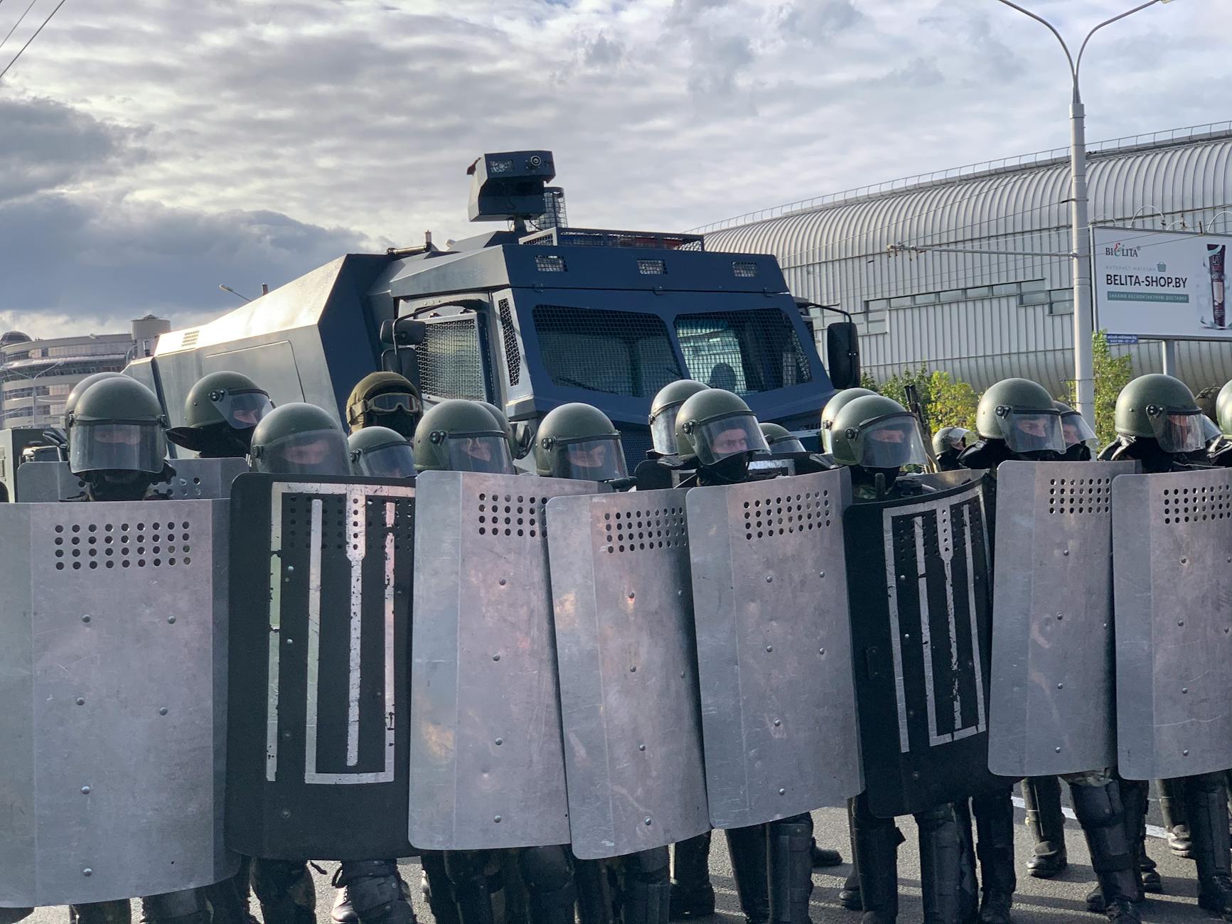 Riot police in protective gear and shields in Minsk, standing in front of an armored vehicle.