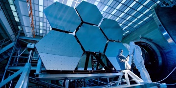 Engineers in protective suits work on telescopic mirrors in a high-tech lab.