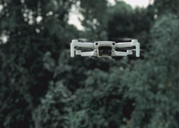 Modern quadcopter with infrared video camera and moving propellers flying under overgrown green trees in daylight