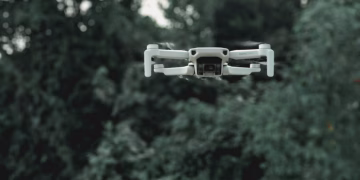 Modern quadcopter with infrared video camera and moving propellers flying under overgrown green trees in daylight
