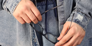 Person in denim outfit holding eyeglasses, showcasing casual fashion and style.