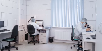 Two laboratory technicians working with computers and microscopes in a clean, modern lab.