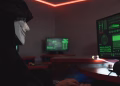 A person wearing a hacker mask operates a computer in a dimly lit room with digital displays.