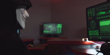 A person wearing a hacker mask operates a computer in a dimly lit room with digital displays.