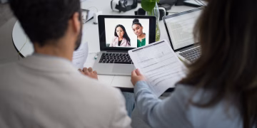 Two professionals conduct a virtual job interview using laptops in a modern office.