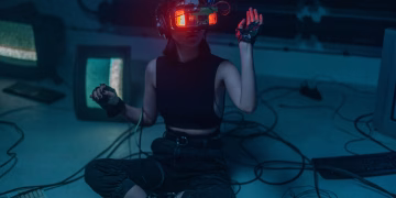 A woman wearing a VR headset sits on the floor surrounded by retro tech in a dark room.
