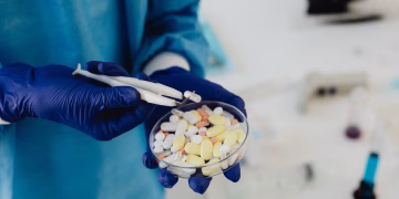 Scientist in gloves using tweezers to handle assorted pills in a petri dish.