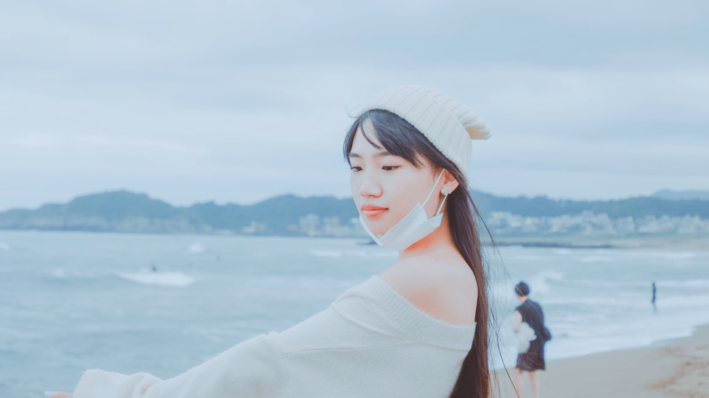 Fashionable woman in a white sweater and beanie by the ocean, wearing a mask.